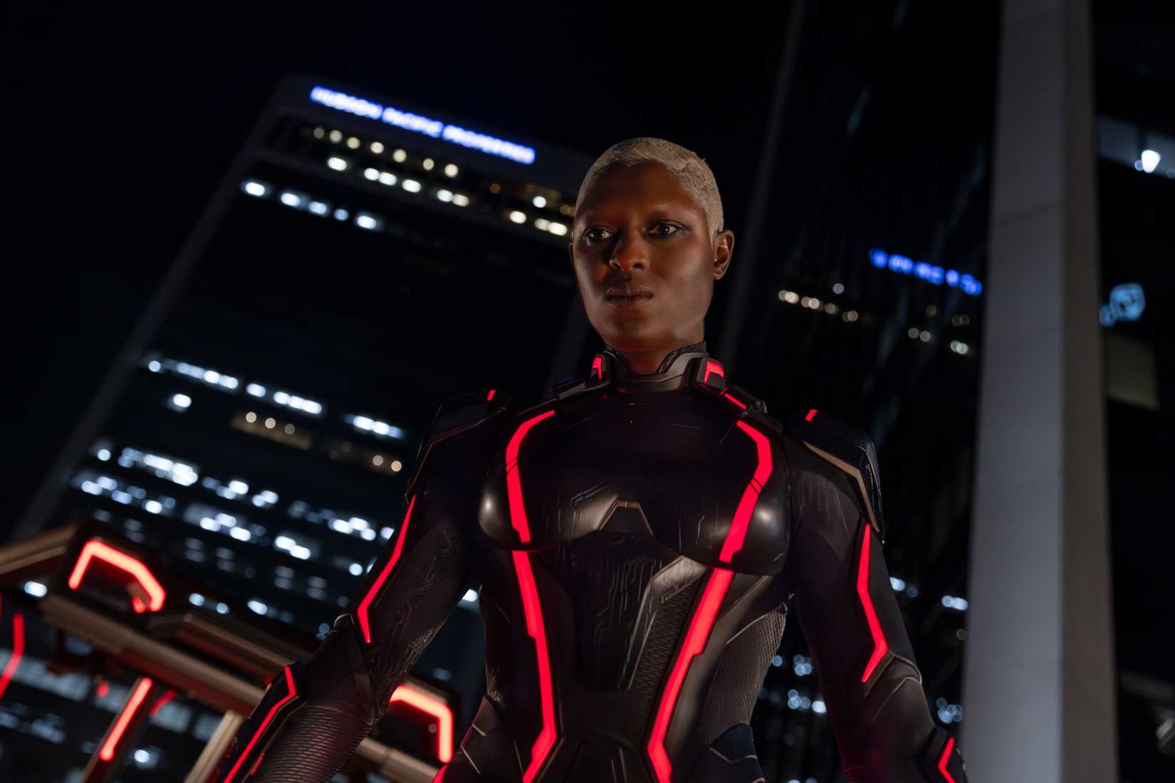 Athena (Jodie Turner-Smith), a Black woman with short-cropped white hair, wearing shiny black armor with glowing red neon highlights, stands in front of a window opening out onto a night cityscape in Tron: Ares